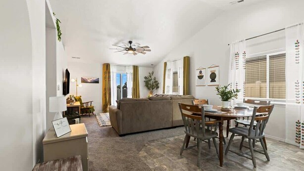 A modern living room with a sofa, dining table, and chairs. There is a ceiling fan, a TV, and a window with curtains. - Retro Caldwell Retreat | Backyard Hammocks | Sleeps 10 (Caldwell)