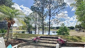 A patio with white chairs, a stone pathway, and a view of a lake with trees and a house in the background. - Magic Sunrise #417 by Shine Villas (Davenport)