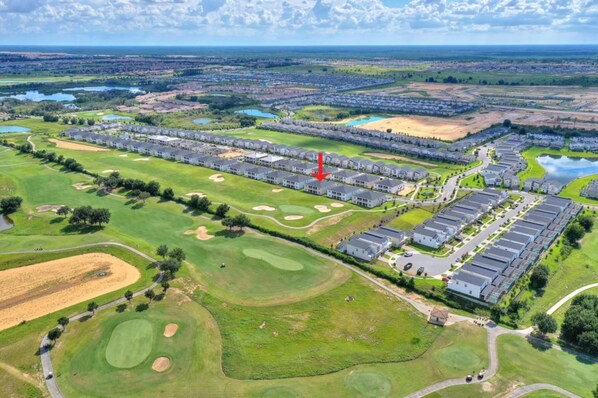 An aerial view of a golf course with residential buildings, including a row of townhouses and a parking area. - Fore Play at Champions Gate #419 by Shine Villas (Davenport)