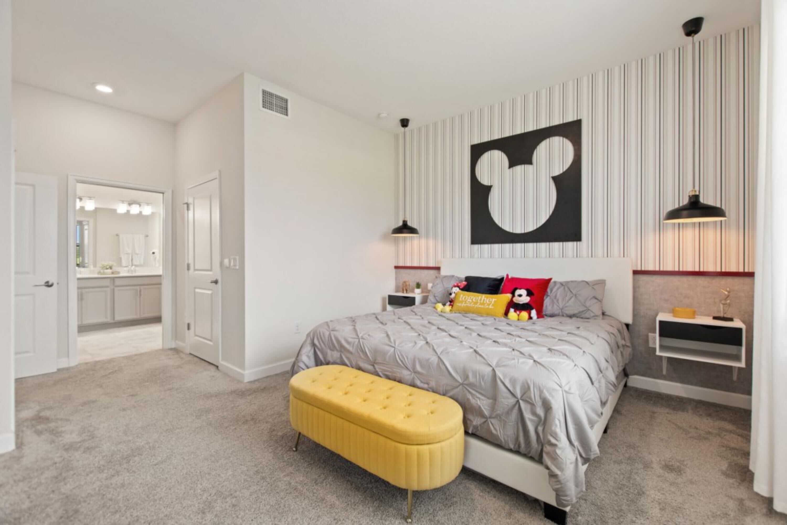 Un dormitorio con cama, un banco, un adorno de Mickey Mouse sujeto a la pared y una mesita de noche con lámpara.