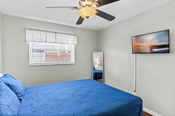 Un dormitorio con cama, ventilador de techo, ventana con persianas, televisor montado en la pared y espejo.