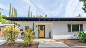 A single-story house with a yellow front door, a small porch, and a garden with palm trees.