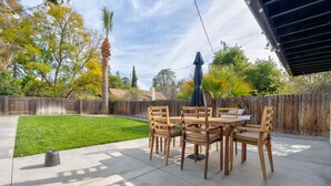 A patio with a wooden table and chairs, surrounded by a wooden fence, with a lawn and trees in the background.
