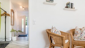 Quarto com cadeiras de vime, planta em vaso, espelho e prateleira com livros.