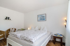 Una cama bien hecha con una colcha estampada, una silla de mimbre, una mesita con libros y una repisa con libros y un cuadro enmarcado.