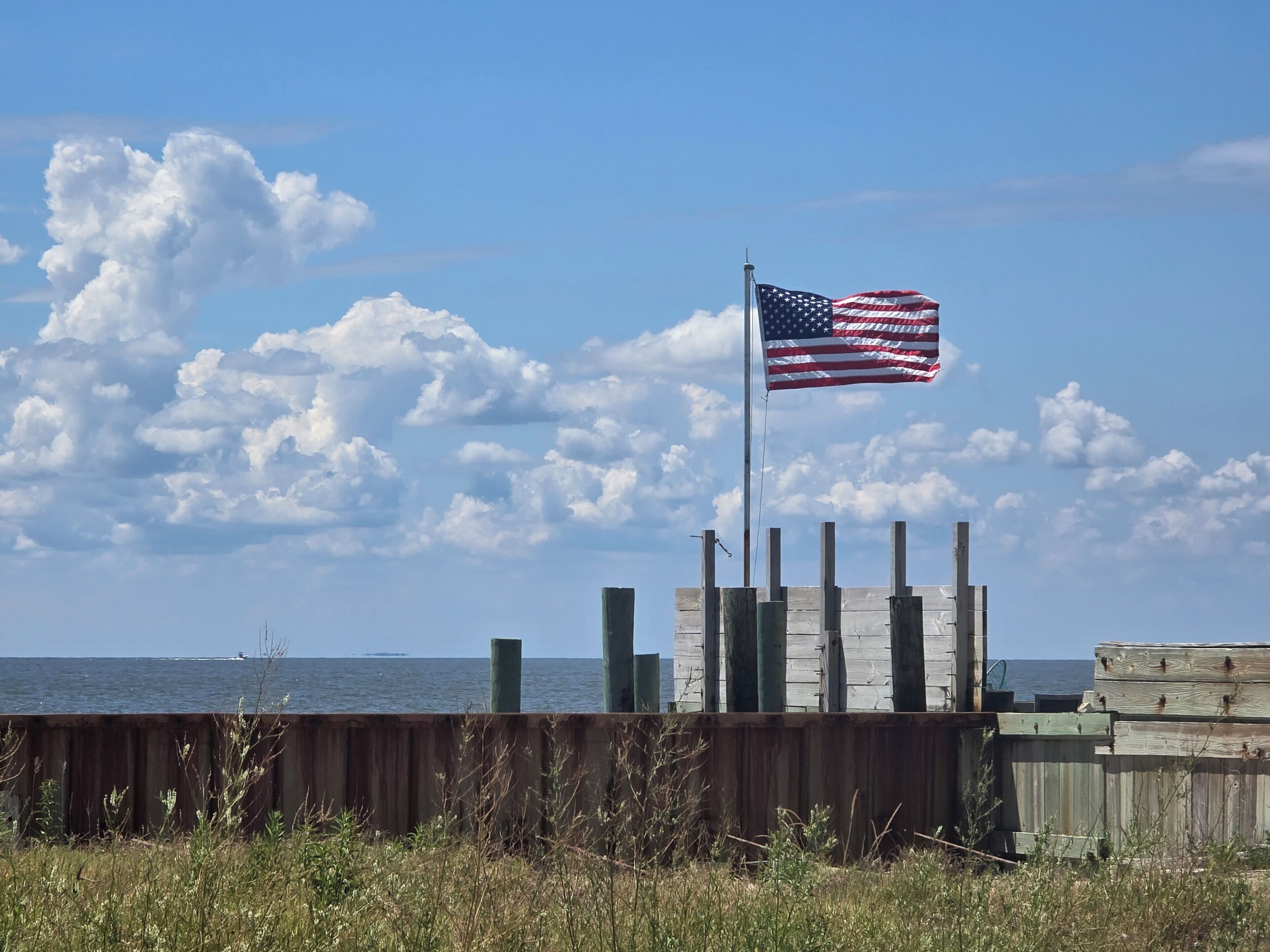 An American flag on a flagpole near a body of water with a wooden fence in the foreground.