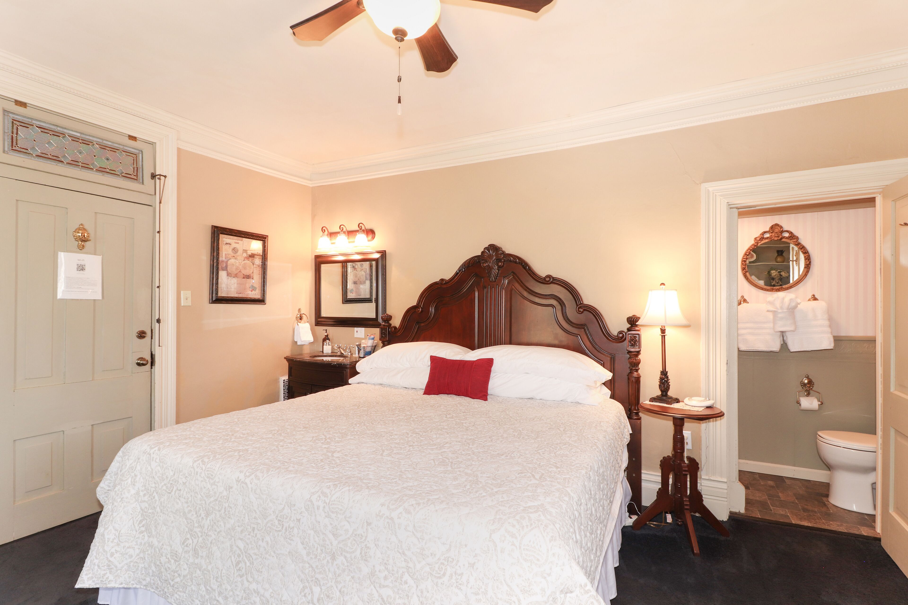A neatly arranged bedroom with a large bed, a bedside table with a lamp, a mirror, and a door with a towel rack.