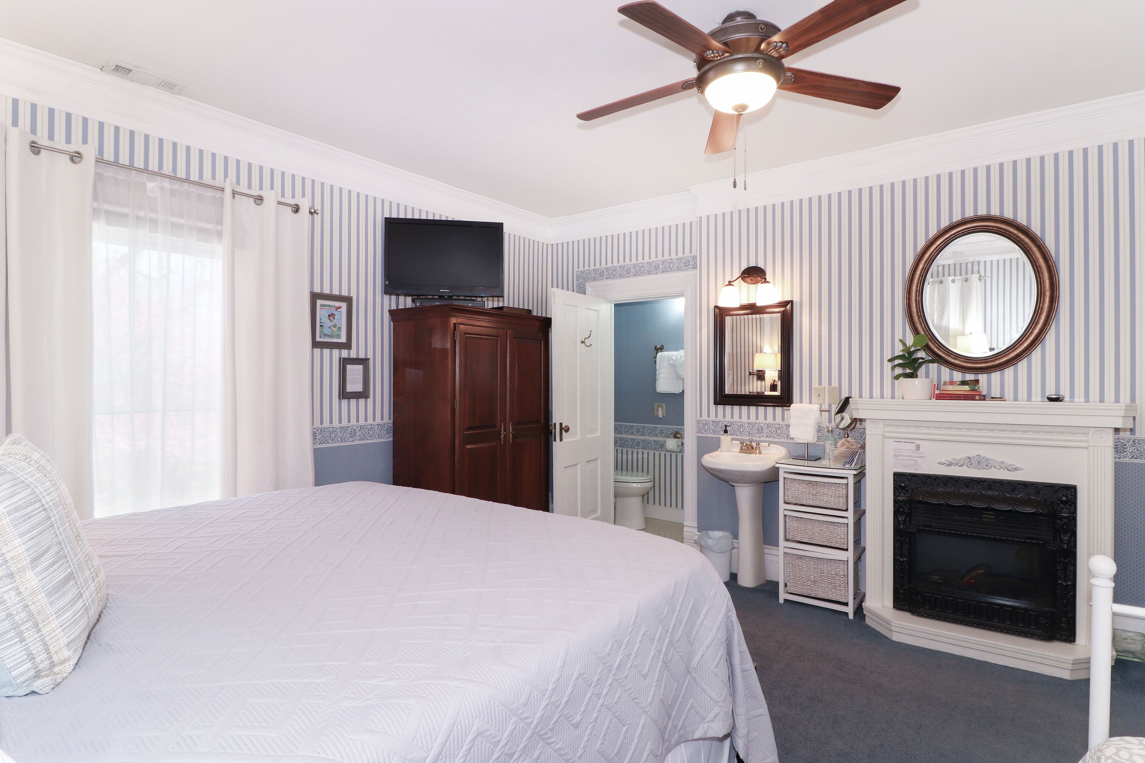 A bedroom with a bed, a ceiling fan, a television, a fireplace, and a bathroom visible through an open door.
