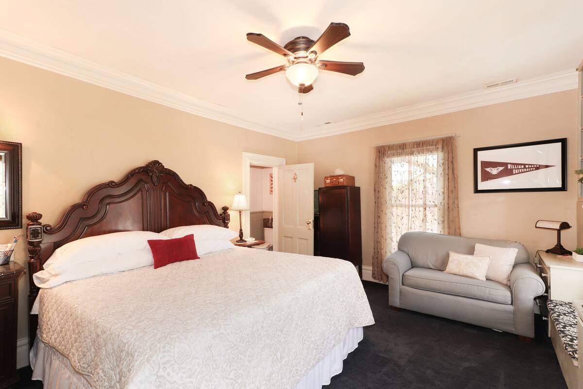 A hotel room with a large bed, a grey sofa, a ceiling fan, and a framed picture on the wall.