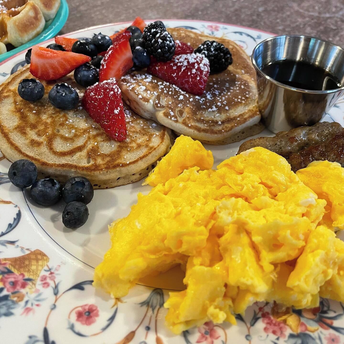 Un plato de panqueques cubiertos de frutos rojos y azúcar glas, acompañado de huevos revueltos y una pequeña taza de metal con jarabe.
