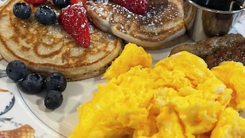 Un plato de panqueques cubiertos de frutos rojos y azúcar glas, acompañado de huevos revueltos y una pequeña taza de metal con jarabe.