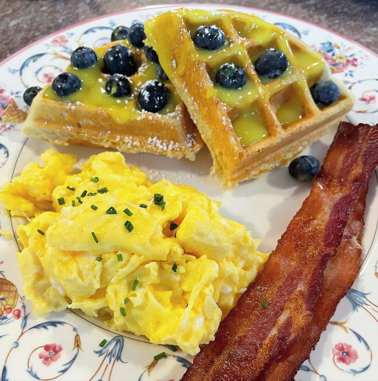 Une assiette avec des gaufres garnies de myrtilles et de sirop, des œufs brouillés et une tranche de bacon.