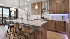 A modern kitchen with wooden cabinets, a large island with bar stools, pendant lighting, and a view through large windows.
