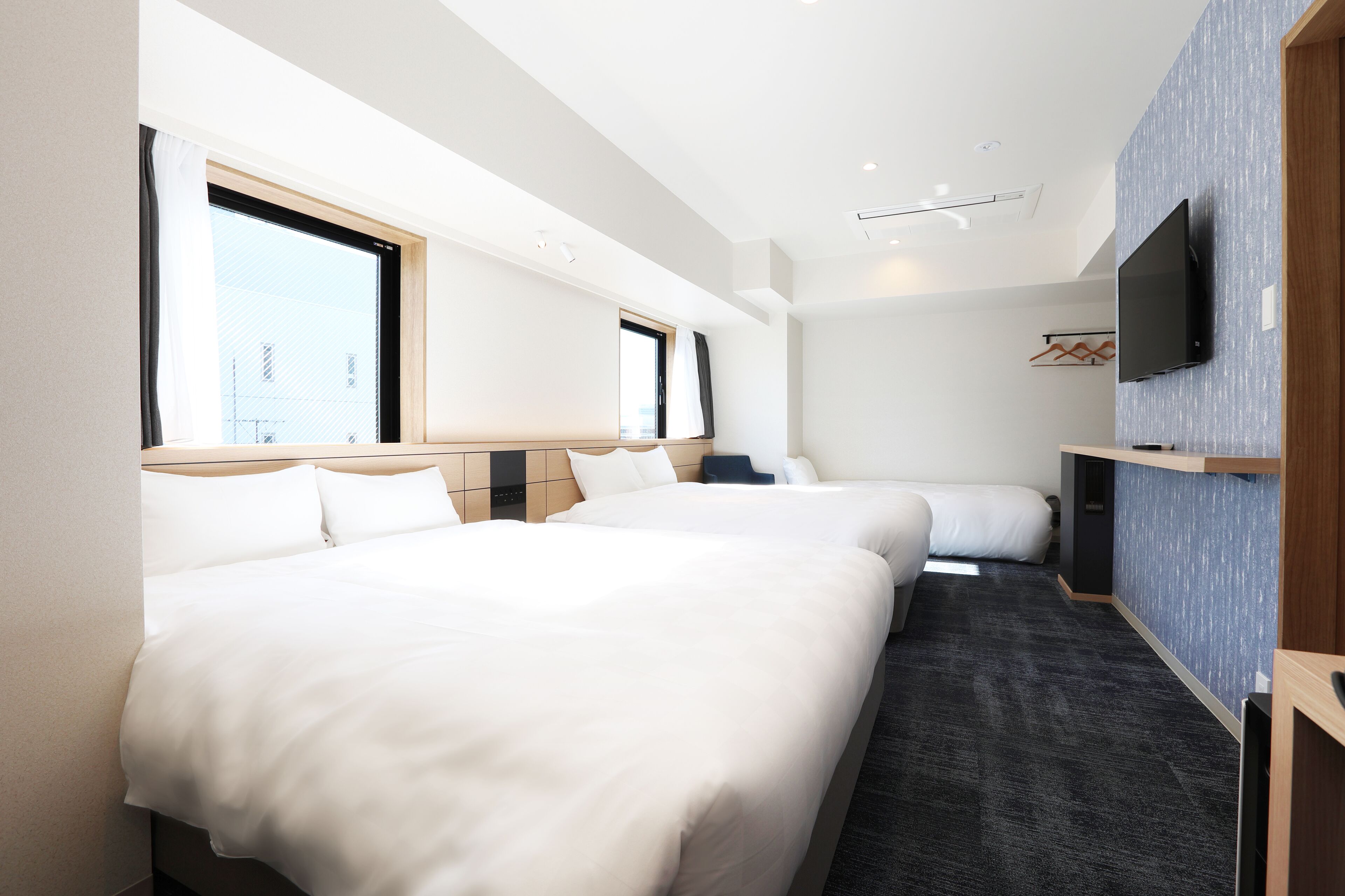 A hotel room with two beds, a flat-screen TV mounted on the wall, and a large window providing natural light.