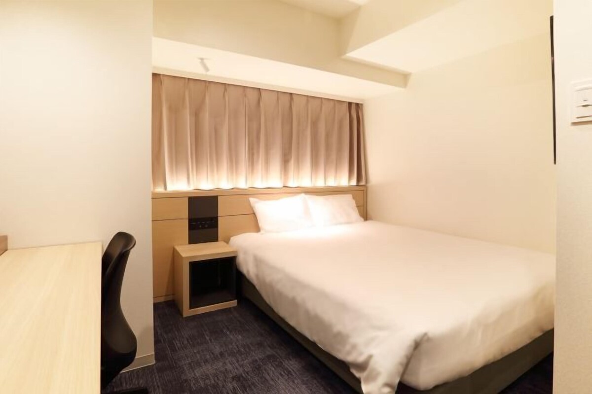 Habitación de hotel pequeña y ordenada con una cama individual, una mesita de noche, una silla y una ventana con cortinas.