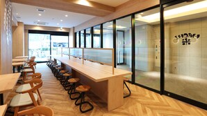 Un restaurante moderno con mesas y sillas de madera, una barra alargada con taburetes y grandes ventanales con vistas al exterior.