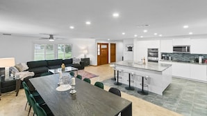 A modern kitchen with white cabinets, a central island with bar stools, and a dining area with a dark wood table and green chairs. - Luxury Villa with Heated Saltwater Pool & Spa, Mini Golf, BBQ, 5 Mins to Beach (Largo)