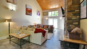 A spacious living room with a high wooden ceiling, a stone fireplace, and a large window allowing natural light. - Blue Mountain Chalet (Bluemountain)
