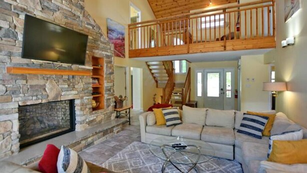 A spacious living room with a stone fireplace, a wooden ceiling, a large flat-screen TV, a sofa with cushions, and a staircase leading to an upper level. - Blue Mountain Chalet (Bluemountain)