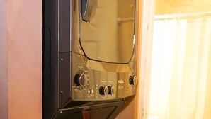 Une machine à laver compacte et moderne, dotée d’un panneau de commande, placée dans un coin d’une pièce dont le sol est carrelé et où l’on aperçoit un rideau au fond.