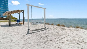 A beach with a swing set, a thatched structure, and a building in the background. - Wake up to Ocean Views from your private balcony! (Tampa)