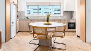 A modern kitchen with a round dining table, two chairs, a built-in oven, and a window with a city view. - Boulevarde 88 - Six (Bondi Beach)