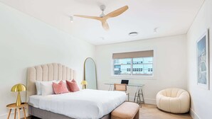 A modern bedroom with a large bed, a desk, a chair, a round ottoman, a ceiling fan, and a window with a view of buildings. - Boulevarde 88 - Six (Bondi Beach)