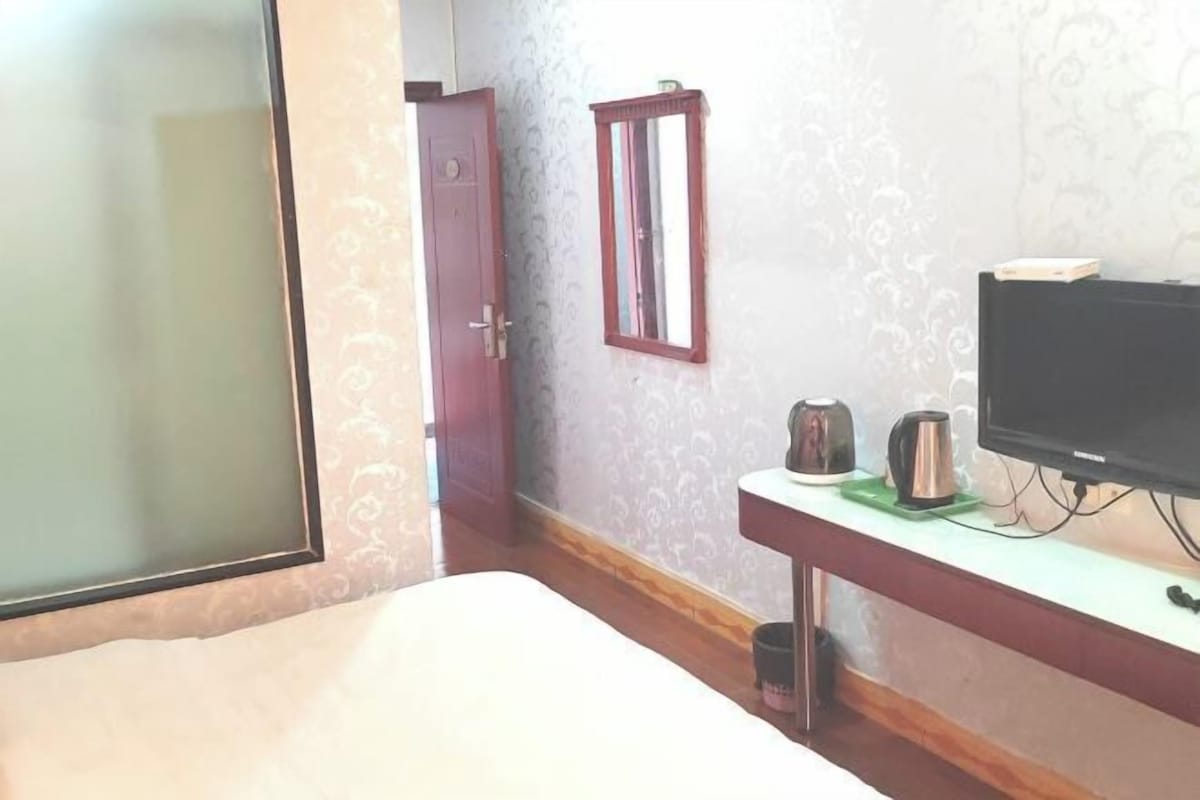 A bedroom with a bed, a television on a stand, a small table with a kettle and a cup, and a door.