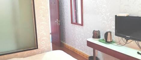 Un dormitorio con una cama, un televisor sobre un mueble bajo, una mesita con un hervidor y una taza, y una puerta.