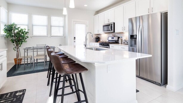 A modern kitchen with white cabinetry, stainless steel appliances, a central island with bar stools, and a dining area with a plant. - Austin Home · Corporate Spacious Luxury Housing (Austin)