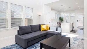 A modern living room with a grey sectional sofa, a black coffee table, and a blue patterned rug. There is a kitchen area with white cabinets and a central island. - Austin Home · Corporate Spacious Luxury Housing (Austin)