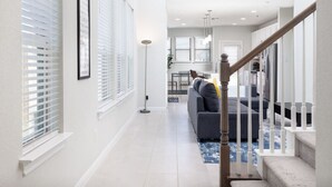 A modern living room with a grey sofa, a wooden staircase, and a large window with blinds. - Austin Home · Corporate Spacious Luxury Housing (Austin)