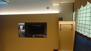 Quarto com televisão na parede, um pequeno armário com objetos e uma janela com vista para o exterior.