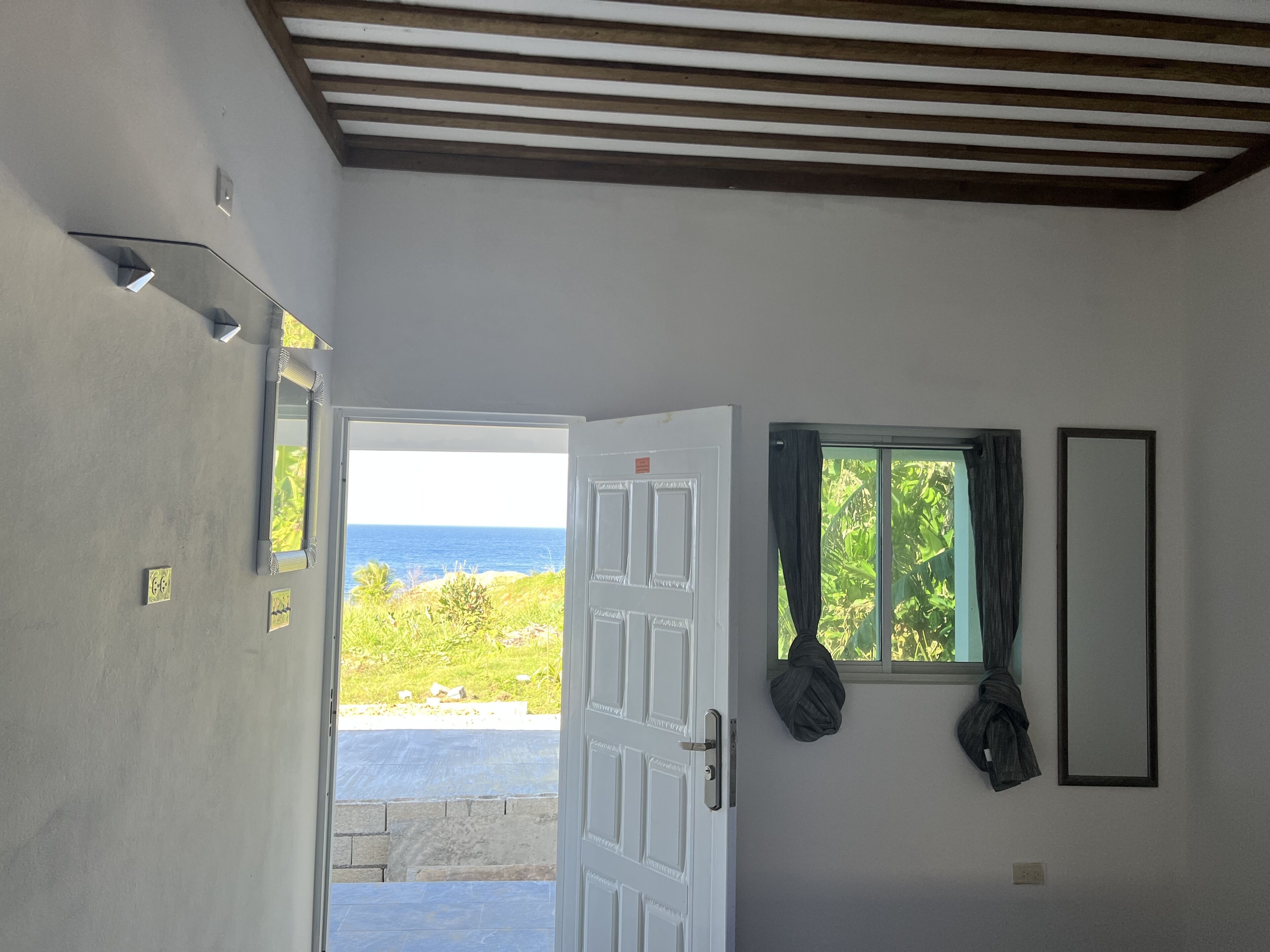 An open door leading to a balcony with a view of the ocean. There is a window with curtains and a mirror on the wall.