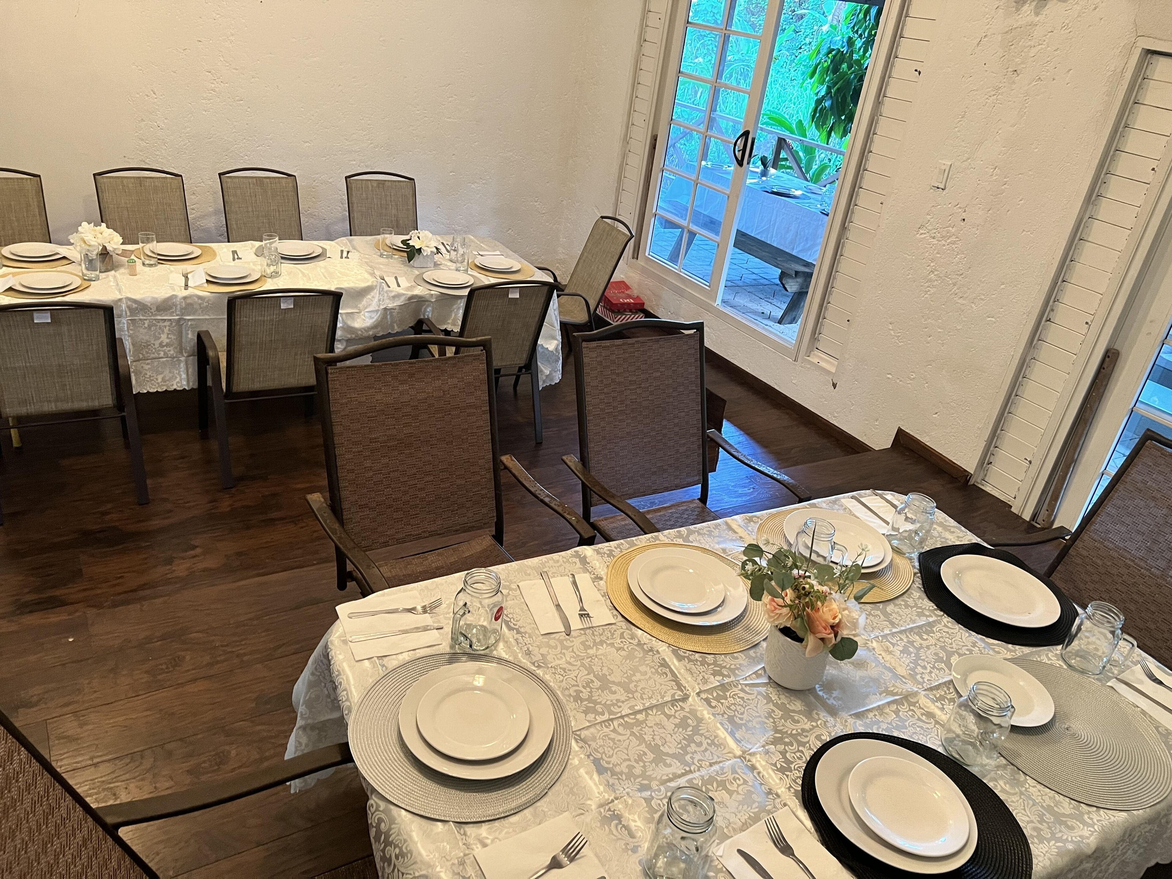 A dining area with a table set for six, featuring a patterned tablecloth, white plates, and dark cutlery. Chairs are upholstered in a light brown fabric.