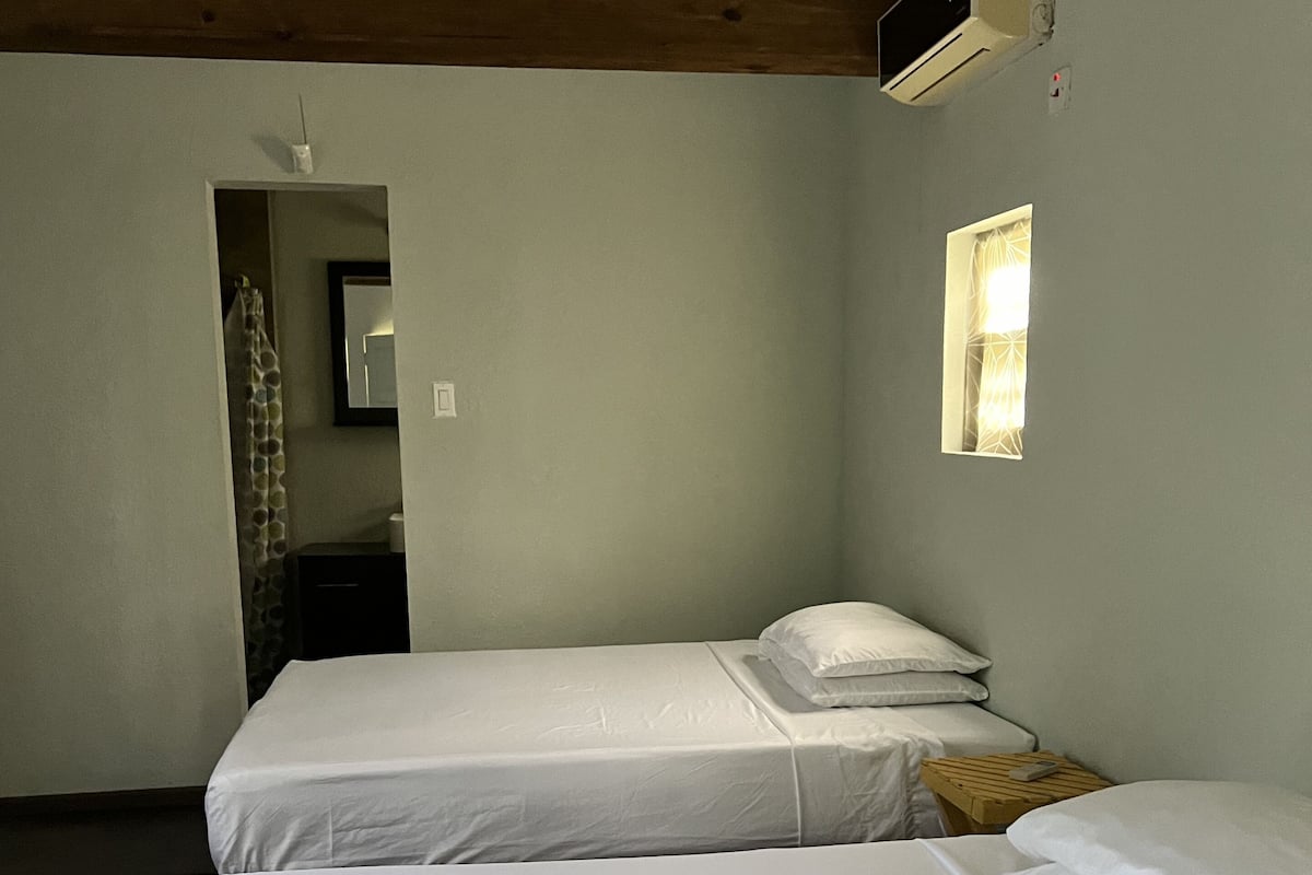 A simple room with two beds, a wooden beam ceiling, a small window, and an air conditioning unit.