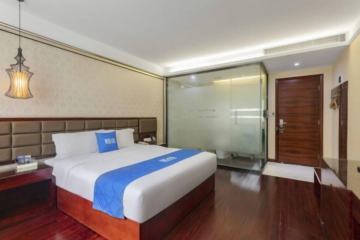 Habitación de hotel con una cama grande, una mesita de noche, un escritorio, una silla, cabina de ducha de vidrio y una puerta de madera.