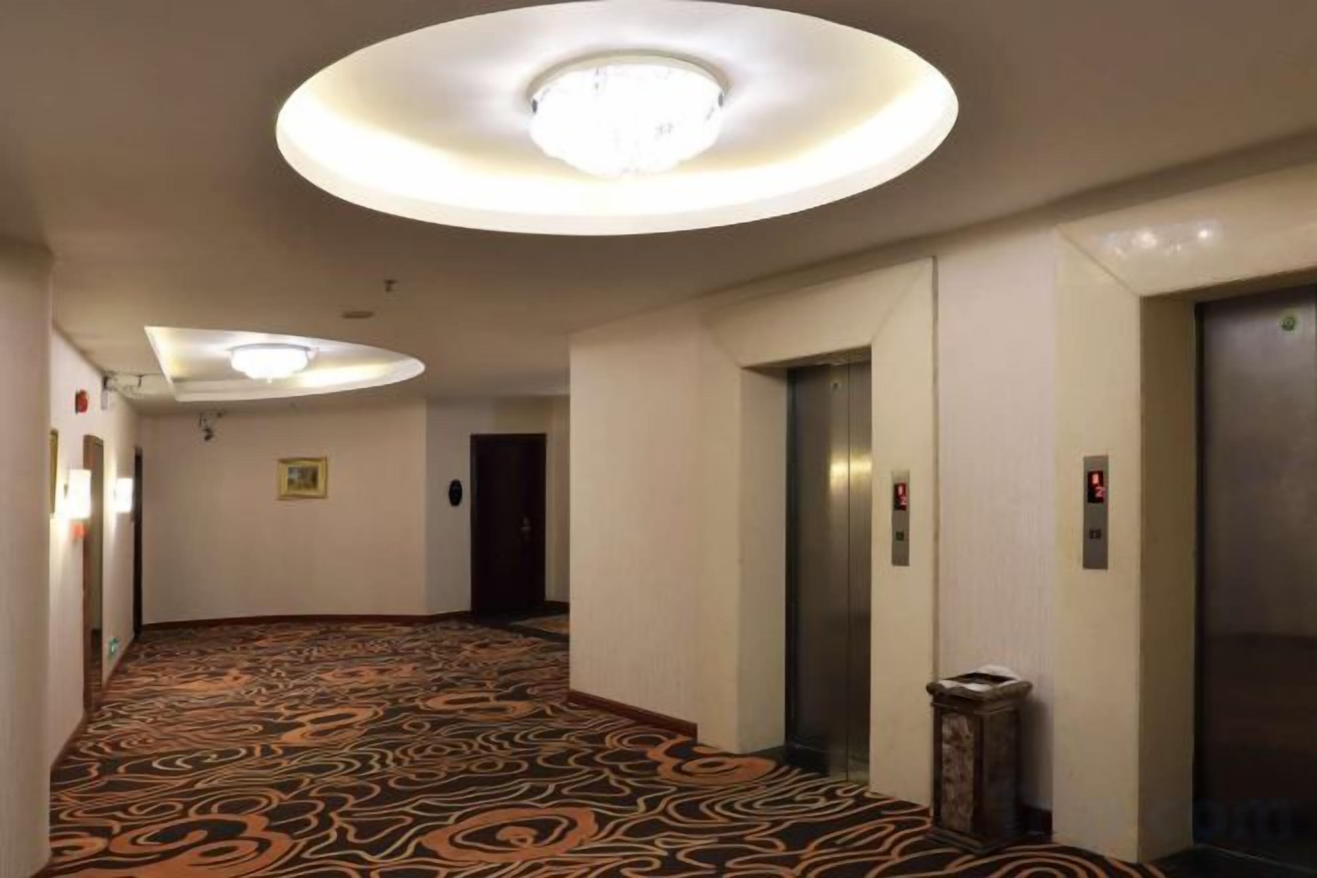 A hotel corridor with a patterned carpet, a circular light fixture, and an elevator.