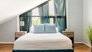 A bedroom with a sloped ceiling, a bed with white bedding and teal accents, two bedside tables with lamps, and a view of greenery outside. - Mt. Adams 1BR Great Location! Steps from Monastery (Cincinnati)