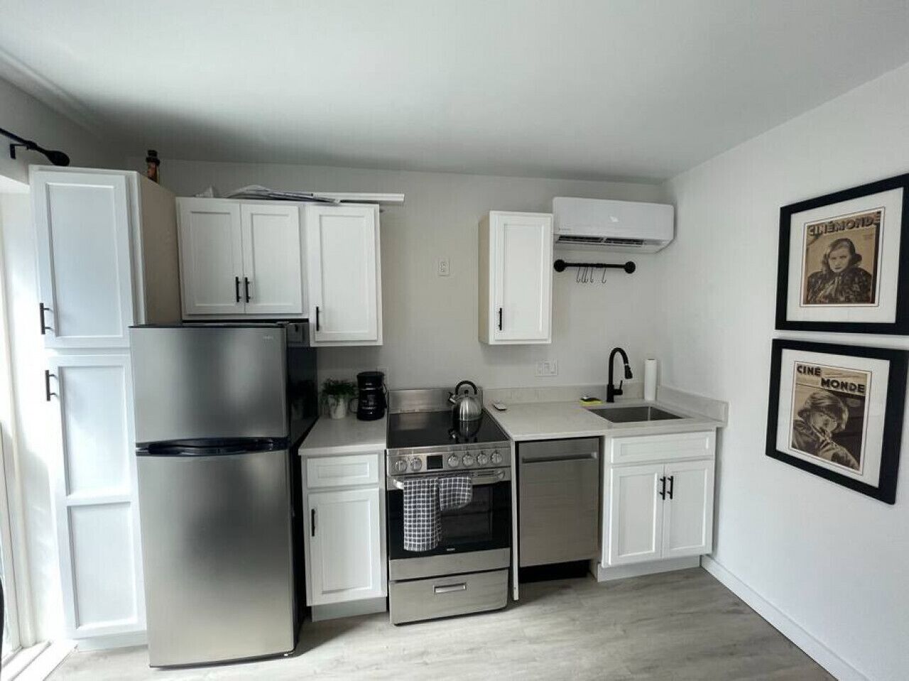 Une cuisine compacte avec des meubles blancs, un réfrigérateur en acier inoxydable, une cuisinière et un lave-vaisselle. Des tableaux encadrés ornent le mur.