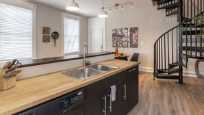 A modern kitchen with a wooden countertop, stainless steel sink, and black cabinetry. There is a staircase with a black railing leading to an upper level.