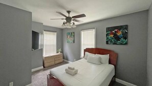 Een slaapkamer met een bed, een plafondventilator, een dressoir, een raam met jaloezieën en een schilderij aan de muur.