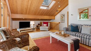 A spacious loft-style living room with a high ceiling, wooden beams, a skylight, a television, a sofa, a striped couch, and wicker furniture. - The Bluebell - Luxury Lodge With Hot Tub (Tenby)