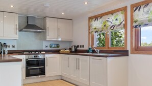 A modern kitchen with white cabinets, a built-in oven, and a window with a floral view. - The Bluebell - Luxury Lodge With Hot Tub (Tenby)
