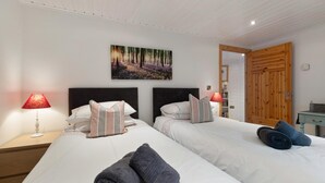A hotel room with two beds, a painting of a forest, a wooden door, and a nightstand with a lamp. - The Bluebell - Luxury Lodge With Hot Tub (Tenby)