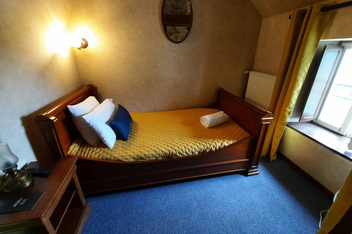 Ein Einzelbett mit Holzrahmen, einem Kopfteil und einer gelben Steppdecke. Ein kleiner Nachttisch mit Lampe und Fernbedienung steht daneben. Ein Fenster mit Vorhängen ist sichtbar, und unter der Wand befindet sich eine Heizung.