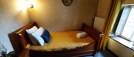 Una cama individual con estructura de madera, cabecera y colcha amarilla. Se observa una mesita de noche con lámpara y control remoto. Se ve una ventana con cortinas, y un radiador en la pared.