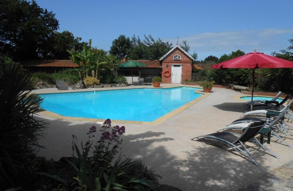 A swimming pool with lounge chairs and a red umbrella, surrounded by greenery and a small brick building.