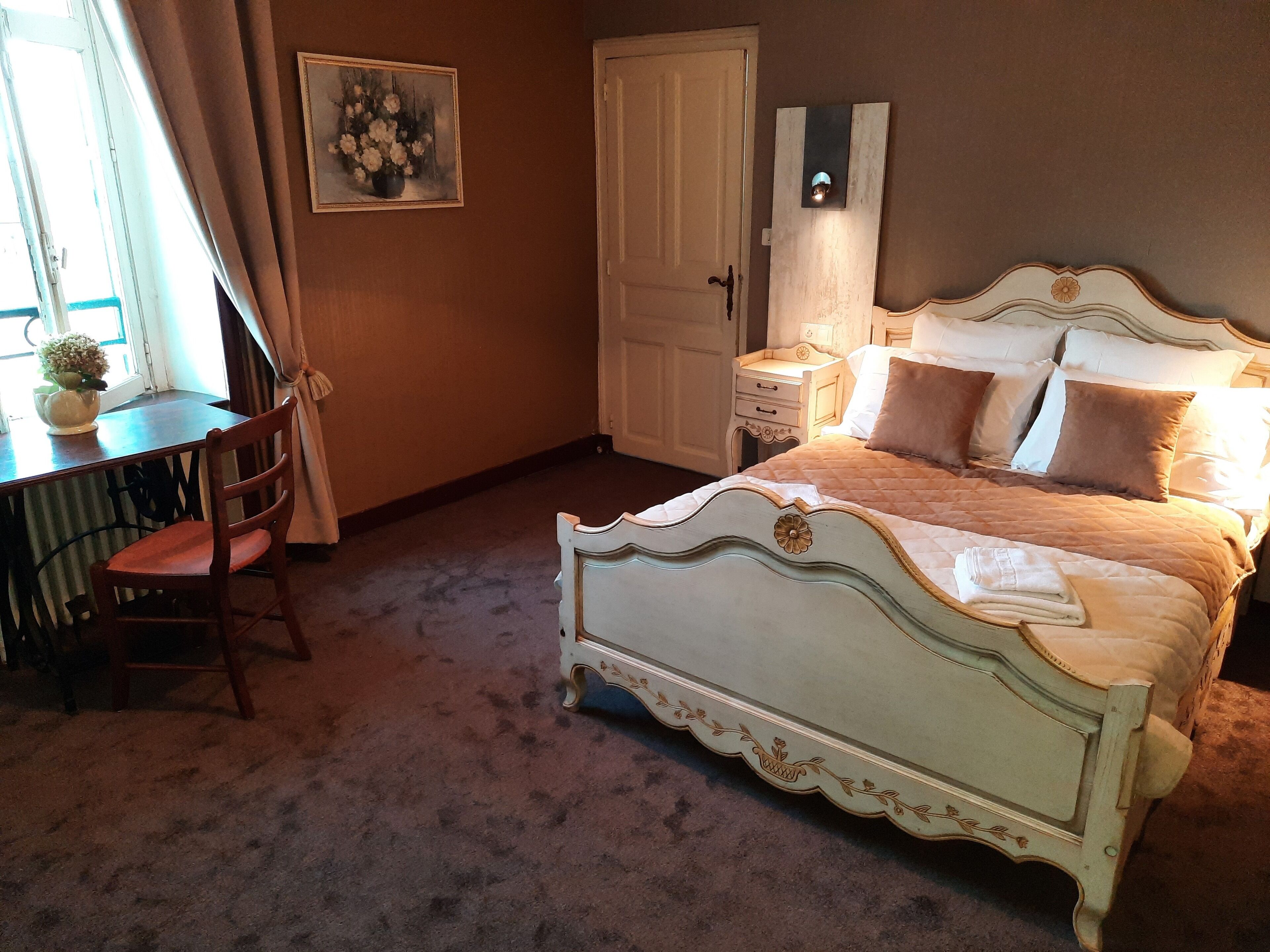 Un dormitorio con cama, una silla, una mesa, una puerta y una lámpara fijada a la pared.