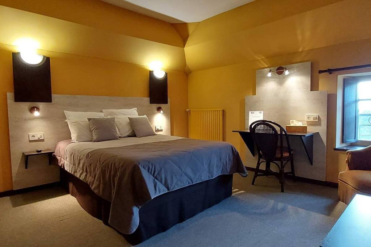 Ein Hotelzimmer mit Bett, Schreibtisch und Stuhl. An den Wänden sind Lampen angebracht, und ein Fenster bietet einen Ausblick.